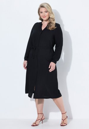 Femme aux cheveux blonds ondulés porte une robe midi noire à manches longues avec des boutons et une ceinture à la taille, associée à des sandales rouges à talons hauts avec brides.