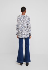 Chemise à manches longues avec un motif paisley bleu et gris sur un tissu blanc, associée à un pantalon en denim évasé. L'ensemble présente une coupe décontractée.