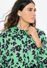 Camicia verde con bottoni a pressione, caratterizzata da un motivo astratto nero, con colletto classico e bottoni verdi, indossata da un modello con capelli lunghi e mossi.