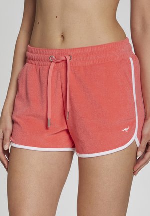 Nahaufnahme einer Person, die korallfarbene Frottee-Shorts mit weißem Rand und Kordelzug in der Taille trägt, mittleren Bereich und Hände an den Seiten zeigt.