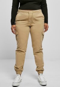 Beige cargo pants with elastic cuffs, drawstring waist, and side pockets, paired with white sneakers. Fabric appears smooth and durable.