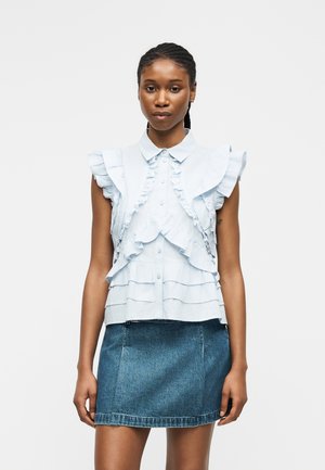 Femme portant un chemisier sans manches bleu clair à volants et une mini-jupe en jean bleu, debout devant un fond gris uni.