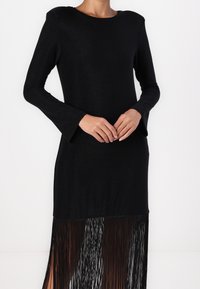 Abito lungo nero a maniche lunghe con scollatura rotonda, caratterizzato da un orlo svasato decorato con frange nere, tessuto testurizzato e silhouette slanciata.