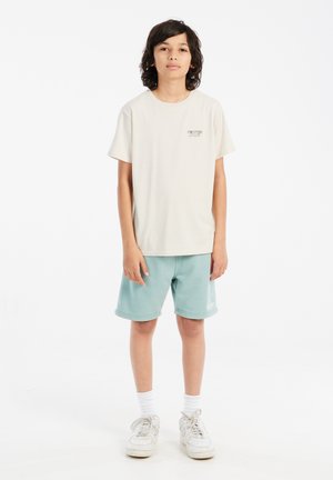 Helle beige Baumwoll-T-Shirt mit "PROTEST"-Logo auf der Brust, kombiniert mit türkisen Shorts, weißen Socken und weißen Sneakers.