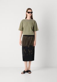 T-shirt di cotone verde oliva con maniche corte, abbinato a una gonna nera plissettata con un motivo marrone. Sandali a slide neri con tacco completano il look.