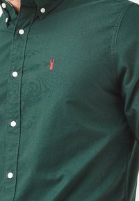 Zielona koszula z guzikami, wykonana z bawełny o gładkiej teksturze, z małym haftem czerwonego homara na lewej piersi oraz białymi guzikami.