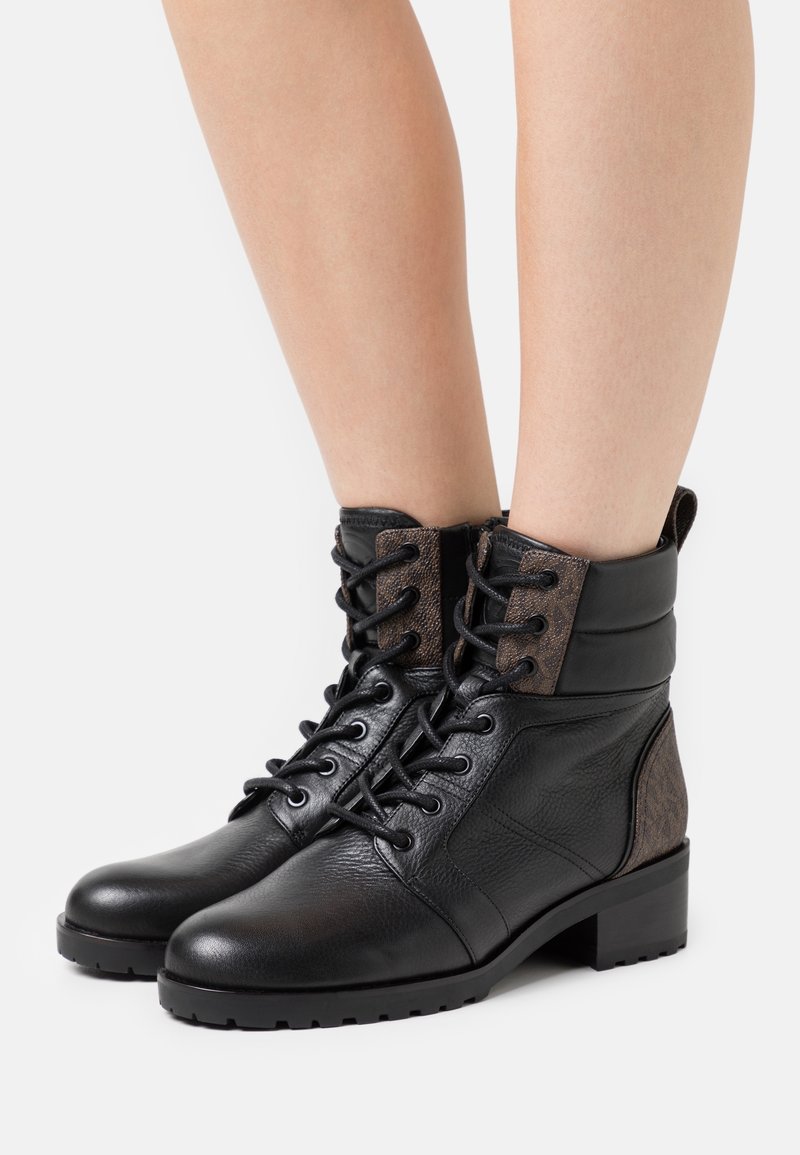 Bottines en cuir noir avec lacets sur le devant, accents en tissu marron texturé sur la languette et le talon, portées sur des jambes nues sur fond blanc.