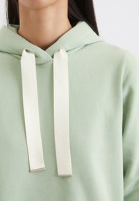 Lichtgroene hoodie met een zachte textuur, met een trekkoord met crèmekleurige uiteinden en subtiele merkvermelding op het koord.