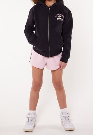 Enfant portant un sweat à capuche noir zippé avec l'inscription « Santa Monica Beach Club », un short rose clair, des chaussettes blanches et des baskets blanches.