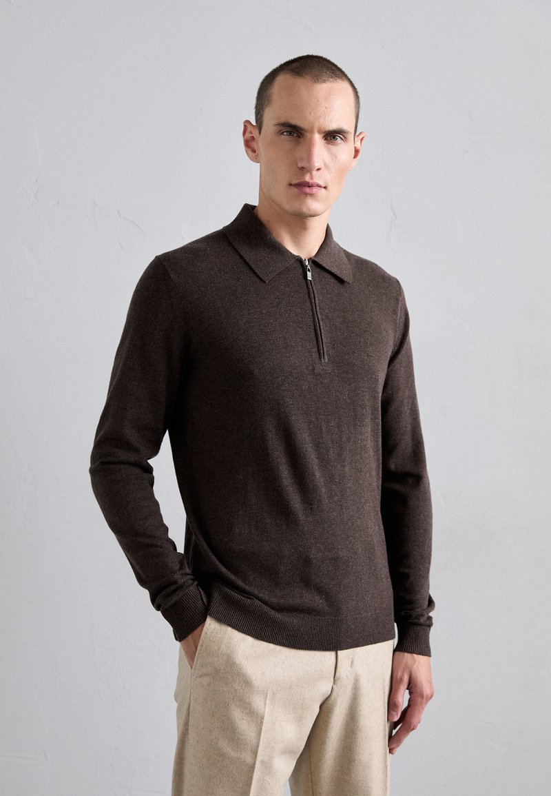 Suéter de polo de manga larga marrón con cuello con cremallera, puños acanalados y dobladillo. Lleva puesto un pantalón beige de corte recto, con textura suave.