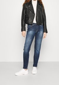 Černá kožená motorkářská bunda, krémový rolák, modré slim-fit džíny a bílé tenisky. Bunda má zipy a zkrácený design.