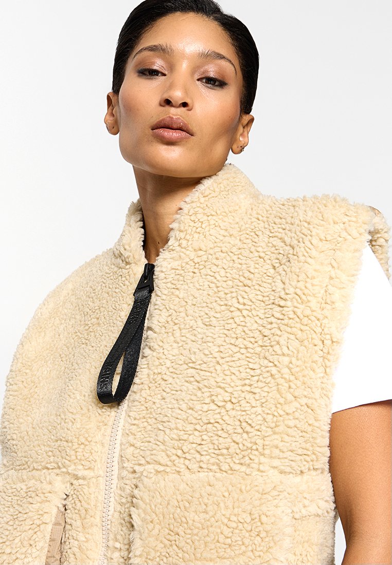 Gilet in finta pelliccia beige con colletto alto, chiusura con zip e cinturino nero a contrasto. Presenta una superficie testurizzata e morbida e un design senza maniche.