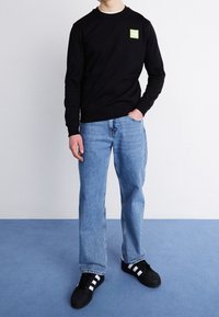 Persona con sudadera negra con logo verde, jeans claros azules y zapatillas negras con rayas blancas, de pie sobre un suelo azul.