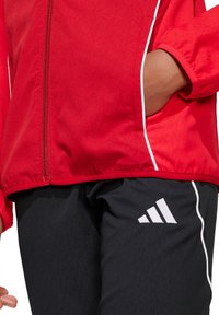 Rode jas met een rits en witte accenten, gecombineerd met zwarte broek met een wit Adidas-logo en zijstrepen.