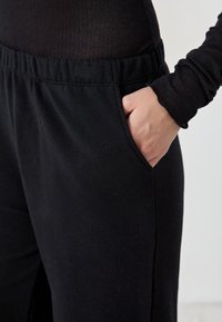 Schwarze, dehnbare Jogginghosen aus Stoff mit einem breiten elastischen Bund und Seitentasche. Die Textur erscheint glatt mit leichter Flauschigkeit.