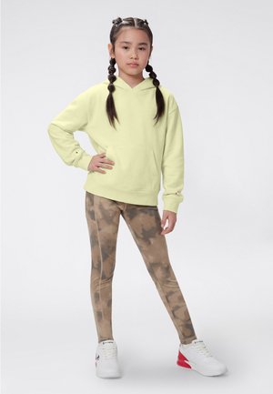 Ragazza con i capelli intrecciati, in piedi con la mano sui fianchi, indossa una felpa con cappuccio giallo chiaro, leggings marrone con effetto tie-dye e sneakers bianche con dettagli rossi.