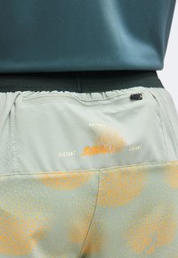 Κοντινό πλάνο της τσέπης από αθλητικά σορτς ανοιχτοπράσινου και πορτοκαλί χρώματος με φερμουάρ και το κείμενο "Distant Dreams" πάνω από μαύρη μέση.