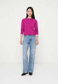 Magenta sweter dzianinowy z długimi rękawami, okrągłym dekoltem, w zestawieniu z jasnoniebieskimi, przetartymi spodniami o szerokich nogawkach i czarnymi butami. Proste tło.