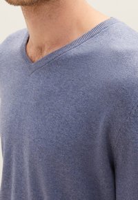 Maglione a V in morbido tessuto a maglia blu chiaro, con una sottile trama e un piccolo logo sul petto. Dettaglio della scollatura e della spalla.