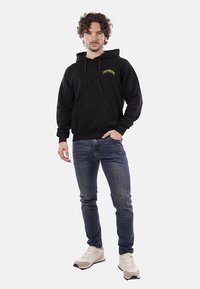 Felpa nera in cotone con tasca frontale e logo in testo giallo, abbinata a jeans blu di denim e sneakers chiare.