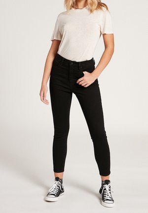 Kvinde iført en ensfarvet hvid t-shirt, sorte high-waisted skinny jeans og sorte høje sneakers med hvide snørebånd, står foran en ensartet baggrund.