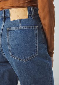 Vue arrière d'une personne portant un jean en denim bleu avec une étiquette en cuir FILIPPA K et un haut à manches longues marron à côtes.