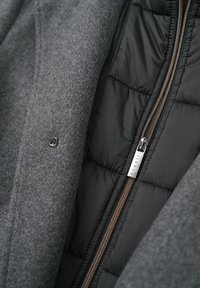 Grau-wollige Außenschicht einer Jacke, mit einem weichen, schwarzen gepolsterten Innenfutter und einem Reißverschluss in Bronzeoptik. Enthält ein kleines Stoffetikett im Inneren.