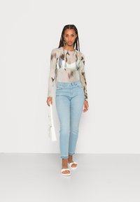 Camisa de manga longa com padrões abstratos em bege e verde, combinada com jeans skinny azul claro e sandálias brancas. Segura uma pequena mala branca.