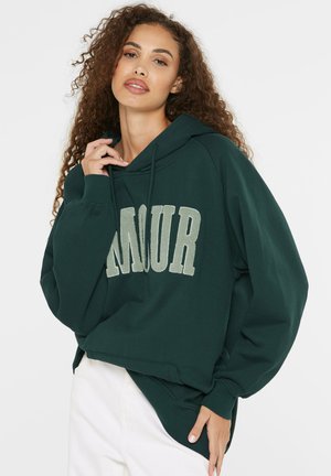 Woman with curly hair wearing dark green hooded sweatshirt with "AMOUR" text and white pants, posing with hand near neck and waist.