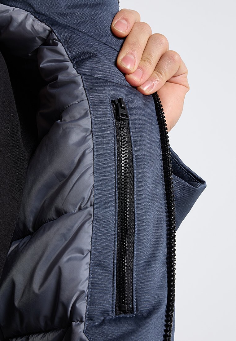 Marineblauwe jas met een gewatteerde voering, voorzien van een zwarte ritszak en een gladde textuur. De stof lijkt waterafstotend te zijn.