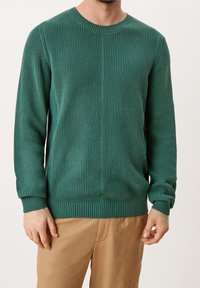 Homme portant un pull côtelé turquoise et un pantalon beige, debout devant un fond clair uni, les mains détendues le long du corps.