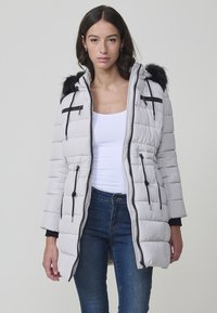 Mujer que lleva una chaqueta acolchada gris claro con capucha de piel negra, cremalleras negras y cordones, combinada con una blusa blanca y jeans azules.