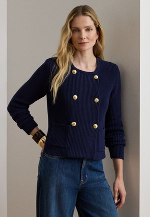 Pikkade blondide juustega naine, kellel on seljas tumesinine kootud kardigan kuldsete nööpidega ja sinised teksad, seisab ühe käega puusal.