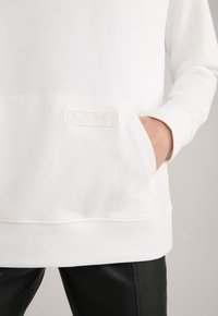 Sweat-shirt blanc à manches longues avec une poche avant, main dans la poche, et un petit logo "JOOP!" brodé au-dessus de la poche, porté avec un pantalon noir.