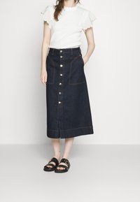 Mörkblå denimkjol med rak silhuett, knappstängning fram, två sidofickor och kontrasterande sömmar. Buren med en vit topp och sandaler.