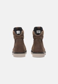 Brązowe skórzane buty typu high-top z czarnymi akcentami, teksturowana powierzchnia, okrągły czubek i gumowa podeszwa, z logo po bokach i metkami wewnątrz.