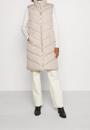 Woman wearing a beige quilted sleeveless long vest with zipper, cream sweater and pants, and black ankle boots, standing against a white wall.