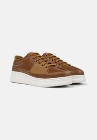 Braune Ledersneaker mit weißen Sohlen, ausgestattet mit Schnürsenkeln und einer strukturierten Oberfläche, in einem Winkel vor einem schlichten Hintergrund präsentiert.