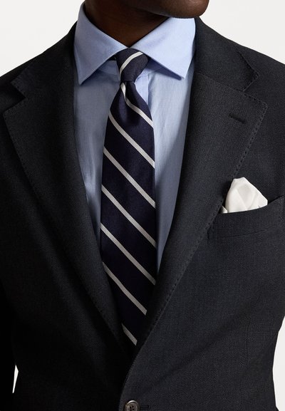 Σκούρο υφασμάτ�ινο σακάκι πάνω από ανοιχτό μπλε πουκάμισο, με ναυτικό γραβάτα με λευκές ρίγες και λευκή μαντήλα τυλιγμένη στο σακάκι.