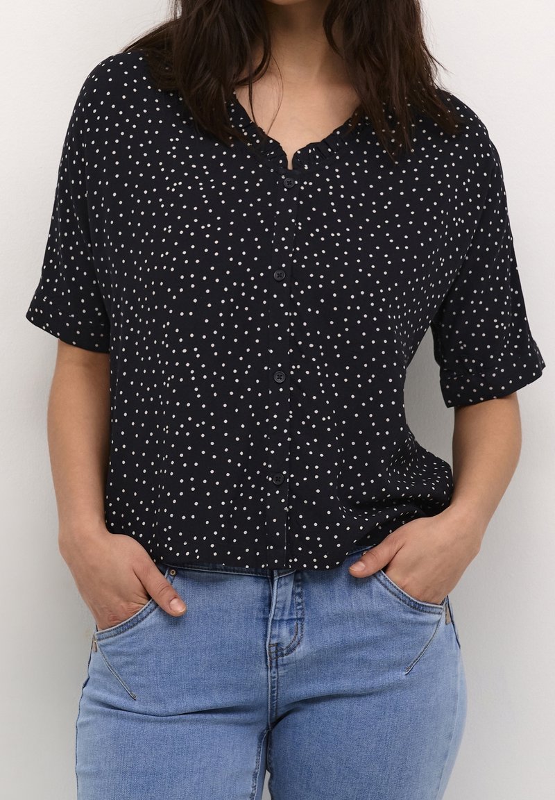 Camicia nera a maniche corte con pois, realizzata in tessuto leggero, con scollo rotondo e vestibilità rilassata, abbinata a jeans in denim blu chiaro.
