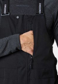 Black waterproof overalls with adjustable straps, featuring a zippered chest pocket. Texture is smooth, made of durable fabric.