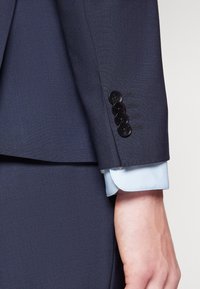 Marinblå kavajärm med fyra svarta knappar, sidstygnsdetalj och ljusblå skjortmanschett synlig under. Slät textur.