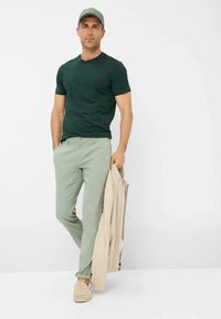 Camicia verde scuro a maniche corte, pantaloni verde chiaro, giacca beige e scarpe slip-on color tortora chiaro. Un berretto grigio completa l'outfit.