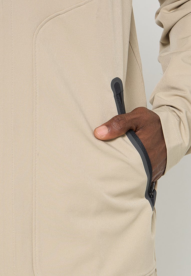 Veste beige au texture lisse ; elle possède une poche zippée avec des accents noirs et des coutures visibles le long des coutures.