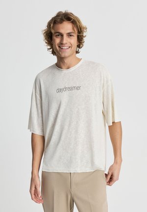 Giovane uomo con capelli ricci che sorride, indossa una maglietta bianca larga con testo "daydreamer" e pantaloni beige, in piedi davanti a uno sfondo uniforme.