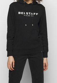 Svart hoodtröja i bomull, med en framlomma, dragskor med metallspetsar och en vit "BELSTAFF"-logotyp tryckt på bröstet.