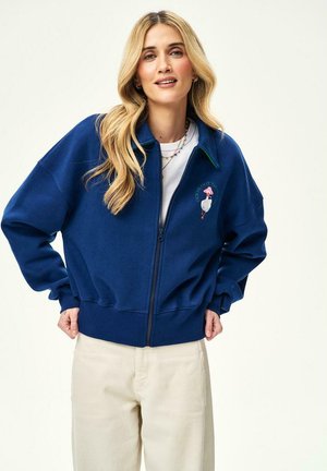 Kobieta z długimi blond włosami, ubrana w niebieską bluzę na zamek z białą grafiką kaczki oraz kremowe spodnie, na jednolitym tle.