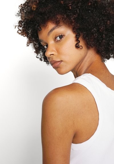 White ribbed tank top showcasing a round neckline, paired with a model with curly hair, displaying textured skin and a natural makeup look.
