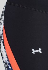 Närbild av svarta träningsleggings med Under Armour-logotyp, med mönstrade sidopaneler i röd och vit.