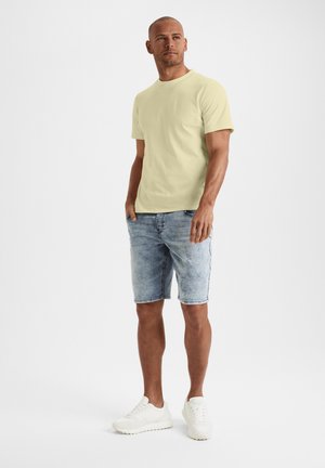 Helles gelbes kurzärmliges T-Shirt, kombiniert mit verwaschenen Jeansshorts und weißen Sneakers. Weicher Stoff, lässige Passform und minimale Details.